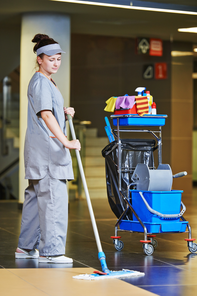 Pracownica sprzątająca myje podłogę mopem obok wózka serwisowego z profesjonalnym sprzętem do sprzątania.