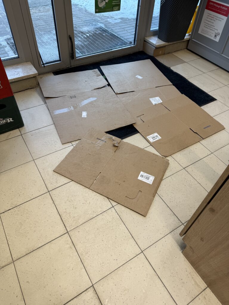 Kartony rozłożone na podłodze przy wejściu do budynku zimą, zabezpieczające przed wilgocią i błotem wnoszonym z zewnątrz.